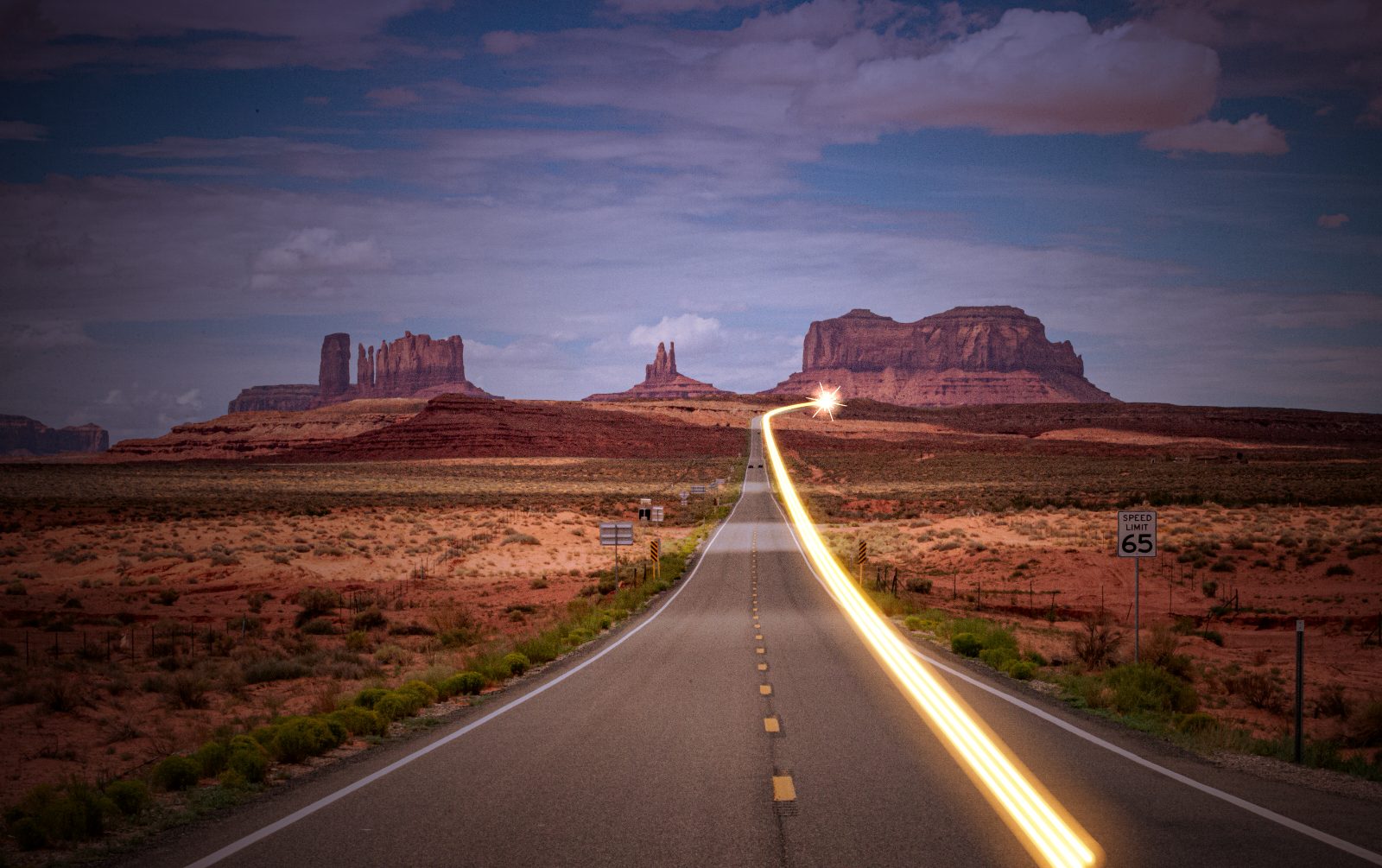 The highway into Monument Valley, Arizona, at dusk, with a glowing light trail tracing the road toward the red buttes.